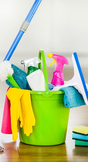 Bucket with cleaning items on blurry modern kitchen background. Washing brush and spray set with copy space.